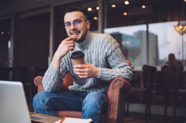 Positive man with beard sitting at table with computer and enjoying coffee while spending time in modern cafe during daytime