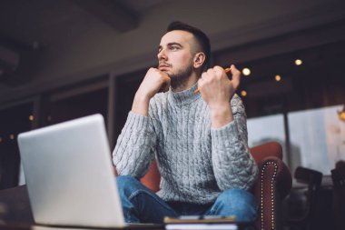 From below of bearded man with pen in hand sitting in soft chair near table with computer and pondering about work