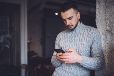 Focused young bearded man in warm knitted sweater standing against wall and using smartphone while spending time in dark room