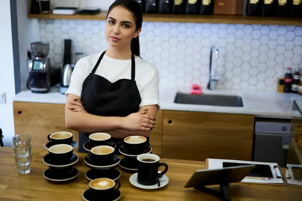 自信に満ちた深刻な女性バリスタ立っていますクロス手で作業場でカプチーノカップ 深刻なウェイトレスでエプロンで注文をする準備ができていますコーヒーハウスのバー — ストック写真