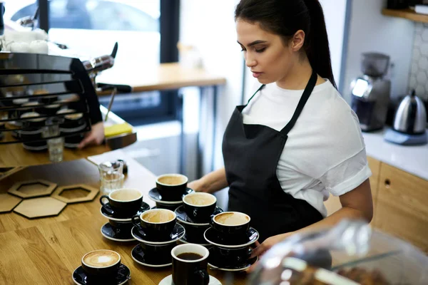 エプロンの熟練女子社員はカフェ内のバーで働くカプチーノマグカップで注文を準備します コーヒーハウスでの仕事に満足プロの女性マネージャー — ストック写真