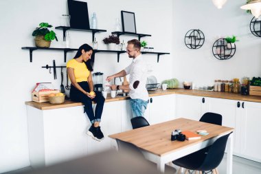 Young man making beverage and talking with female colleague sitting on kitchen table in spacious apartment studio decorated with different decorative items