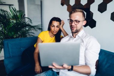 Cheerful man and ethnic woman in casual clothes sitting on sofa and sharing netbook watching video while resting during break at modern office