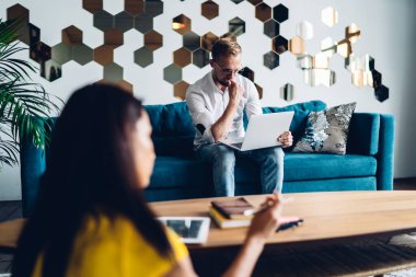 Side view of young bearded man and brunette woman in casual outfit sitting at home office with laptop and discussing business plan for startup