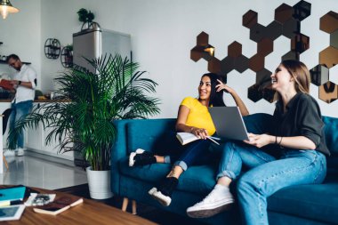Group of cheerful diverse colleagues in casual wear smiling and browsing laptop while working on project together in living room
