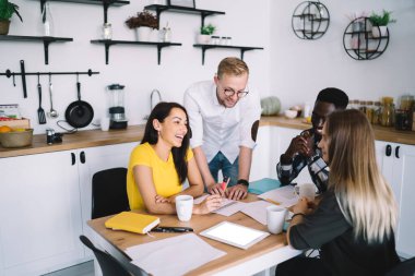 High angle of happy multiracial colleagues in casual outfits discussing business plan while working with papers at wooden table in kitchen