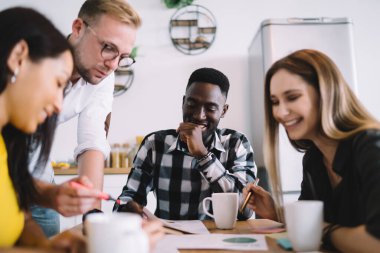 Group of happy multiracial colleagues in casual wear sitting at wooden table and having brainstorm during paperwork together in kitchen
