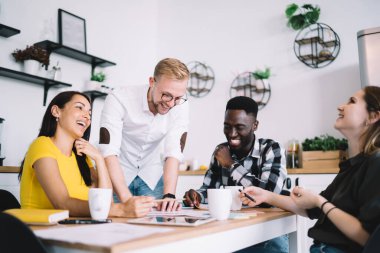 Group of smiling diverse creative colleagues in casual wear having fun while sitting at table and collaborating on task in comfortable home interior