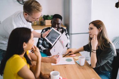 From above of multiethnic young coworkers in casual outfit sitting at table while listening to male colleague using tablet and papers with charts in modern kitchen interior