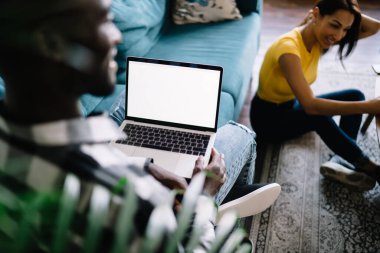 From above of crop African American man using laptop and sitting on sofa while ethnic woman sitting on floor in modern apartment