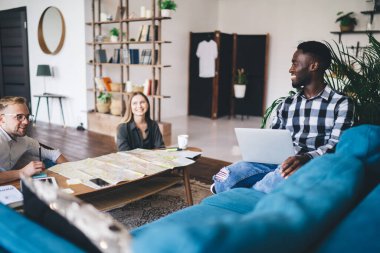 Cheerful young multiethnic friends in trendy clothes sitting near table with gadgets and documents while resting during break in modern workplace