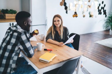 Positive multiethnic coworkers sitting at office table with cups of tea during break and discussing details of project showed on sheet of paper