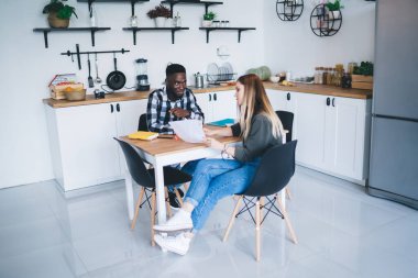 Young African American man and female colleague in casual wear with papers in hand sitting at table in kitchen and working together