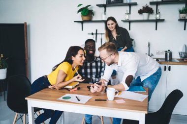 High angle of excited diverse colleagues in casual outfits having conversation about business project while browsing smartphone together during work