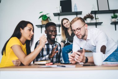 Low angle of male worker showing cellphone to group of positive diverse colleagues in casual clothes during teamwork in creative workplace