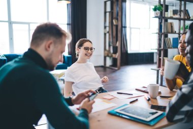 Cheerful young male and female diverse colleagues gathering around table with gadgets and cups of hot drink and discussing business project