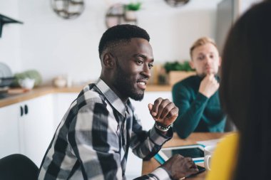 Side view of African American male employee gathering around table with colleagues and listening to ideas during brainstorm in modern room