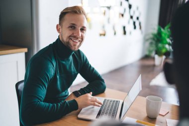 Young handsome businessman in green turtleneck sitting at table with laptop and looking at camera with smile while working on business project in creative workspace shared with other entrepreneurs