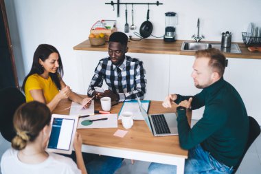 Group of young business people gathering at table and communicating together in kitchen and browsing gadgets while working on project