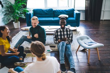 From above of cheerful multiracial partners sitting on rug near papers and copybook during teamwork while looking at each other in flat