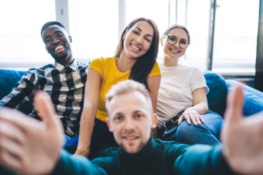 Young content diverse colleagues taking selfie while resting on comfortable sofa near window and looking at camera in flat in sunlight