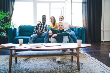 Content young multiracial coworkers in jeans gathering together while sitting on comfortable couch in front of table with cups and papers and looking at camera at home