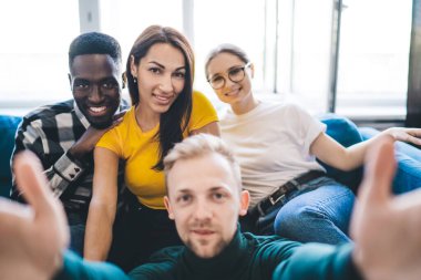 Group of young cheerful multiethnic partners taking self portrait while resting on couch and looking at camera in light apartment