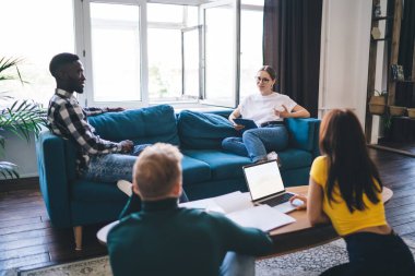 Unrecognizable multiethnic partners with netbook speaking with colleagues on comfortable sofa near window while looking at each other in house