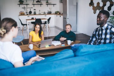 From above of thoughtful coworkers sitting at table with netbook while interacting with multiethnic partners on cozy couch in modern flat