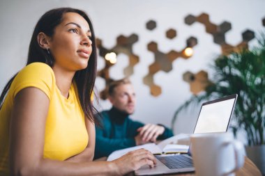 Young ethnic female in bright wear using netbook with empty screen while looking away near male coworker at desk in light workspace