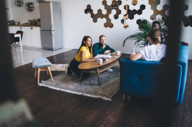 From above of content partners sitting at wooden table with netbook while talking to multiracial colleagues on sofa in modern style apartment with wooden floor