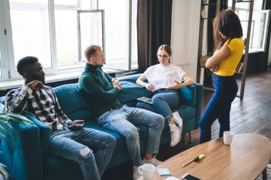 Group of diverse partners on sofa talking to anonymous woman with folded arms while discussing project in light room in flat and looking at each other