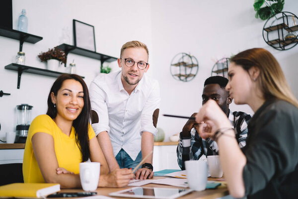 Group of content multiethnic colleagues in casual wear smiling and having discussion about details of business project during meeting in kitchen