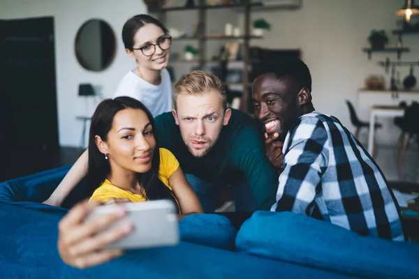 Young cheerful diverse colleagues taking self portrait on cellphone while sitting on cozy couch in living room at home and looking away