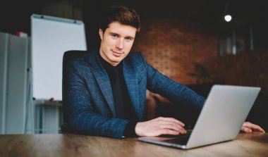 Confident corporate employer using laptop computer during working time in office interior, portrait of Caucasian businessman looking at camera sitting at desktop with modern netbook technology