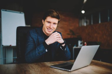 Cheerful corporate employer using laptop computer during working time in office interior, portrait of successful businessman smiling at camera sitting at desktop with modern netbook technology
