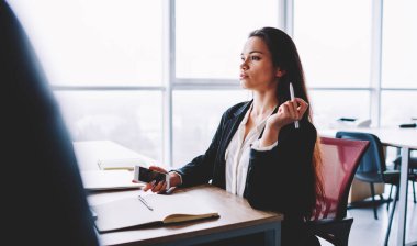 Side view of Caucaisan female entrepreneur with pen and smartphone gadget in hands thinking about business strategy and financial planning, confident secretary in formal wear working at office desktop