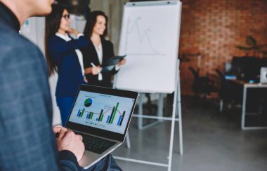 Cropped businessman analyzing financial infographics displayed on modern netbook technology while blurred female colleagues collaborating in office interior, professional browsing graph charts