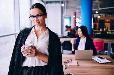 Confident female corporate director in elegant outfit holding coffee cup and looking at camera while employee working on background, portrait of business woman in optical eyewear for vision protection