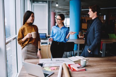 Cheerful male and female colleagues enjoying friendly conversation during coffee break in coworking office, crew of professional architects in smart casual wear communicating about involved project
