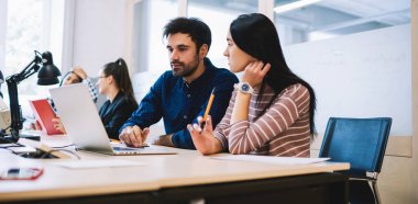 Couple of thoughtful colleagues sitting at table with laptop while working together on new business project in light modern workplace