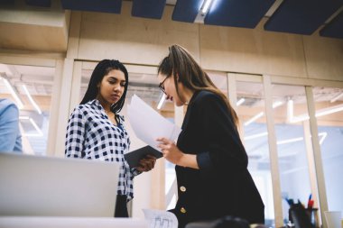 Low angle of diverse pensive female coworkers in casual clothes with tablet and documents exchanging information for business project working in modern office