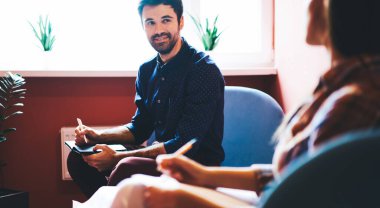 Crop male in casual clothes sitting on chair with pen and notepad while having conversation with female friend in light room