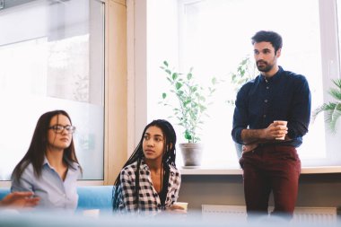 Bearded male standing near window with paper cup of coffee spending time with diverse female coworkers in light modern workplace