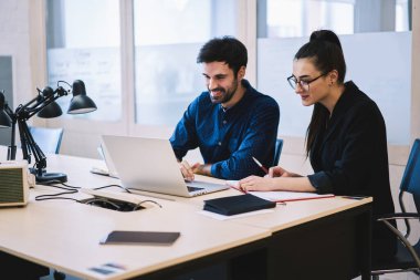 Cheerful young professionals in formal wear checking data on laptop and taking notes in notebook sitting at desk in modern office