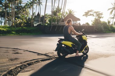 Back view of full body anonymous male driving motorbike and looking away while resting alone in tropical resort and enjoying summer vacation