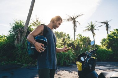 Side view of young bearded man in casual clothes using smartphone while standing on sandy beach with helmet in hand near modern motorbike on sunny day