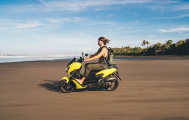 Side view of full length male biker driving electric scooter among sandy beach while looking away and spending summer in tropical country