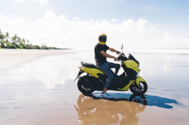 Full body side view of anonymous young male biker in trendy outfit and helmet riding yellow scooter on wet sandy coast near waving ocean on sunny day