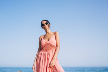 Güneş koruması için güneş gözlüğü takmış kaygısız kadın turistin yarım boy portresi sahilde poz verirken kameraya bakıyor.